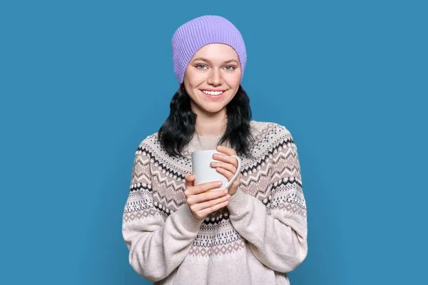 穿着温暖的冬季毛衣的年轻女人手拿着杯子戴着蓝色工作室背景的帽子微笑 年轻快乐的女性 带着牙齿的微笑 过冬的时候 新年的概念 — 图库照片