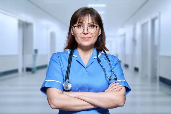 Portrait of confident female doctor in hospital. Middle aged medic with stethoscope with arms crossed in clinic corridor. Medicine, staff, health care, health service, nhs concept