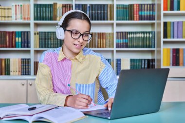 Kütüphanede okuyan genç kız öğrenci. Kulaklıklı bir kadın dizüstü bilgisayar kullanıyor, bilgisayara bakıyor, webinar izliyor, not defterine not alıyor. Ergenlik, teknoloji, eğitim, lise konsepti