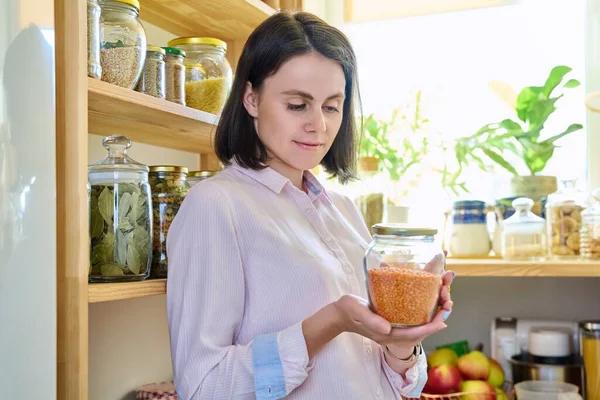 Young woman in kitchen with containers jars of cereals, pasta, dry fruits, holding a jar of red lentils. Female in home pantry, food organization and storage, kitchen utensils, household