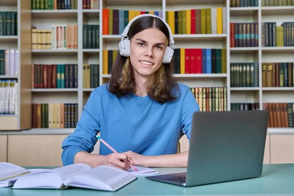 Portrait of teenage student guy in headphones sitting at desk in library, using laptop for studying, taking notes. Smiling young male looking at camera. Youth technology education college university