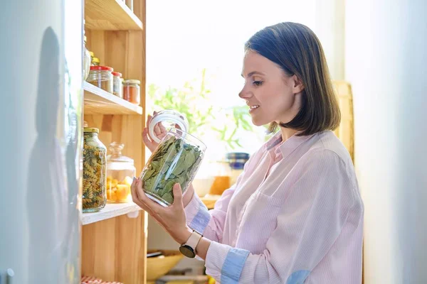 厨房里的年轻女人 她端着盛满谷物 干果的容器 手里拿着一罐干月桂叶 家庭厨房 食品组织和储存 厨房用具 家庭中的女性 — 图库照片
