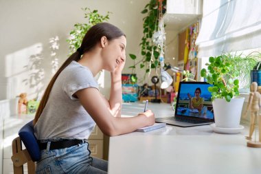 İnternetten ders alan ergen kız dizüstü bilgisayara bakıyor, öğretmeniyle konuşuyor ve not defterine not tutuyor. Uzak sınıf çocukluk çağı e-öğrenim e-eğitim, eğitim teknolojisi