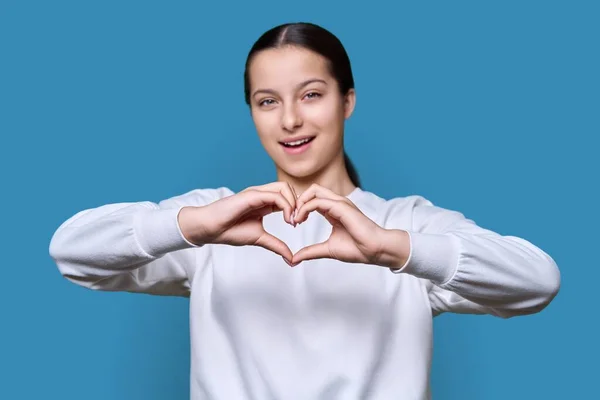Young teenage female showing heart gesture with hands, on blue color background, face out of focus. Adolescence, youth, lifestyle, high school, emotions concept