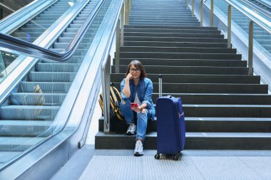 İstasyonda bagajı olan kadın turist. Bavul akıllı telefonlu orta yaşlı bir kadın tren istasyonunun merdivenlerinde oturuyor. Seyahat bagajı insan teknolojisi kavramı