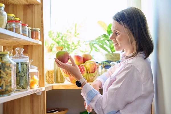 Young woman in kitchen with containers jars of cereals, pasta, dry fruits, holding red apple. Female in home pantry, food organization and storage, kitchen utensils, household