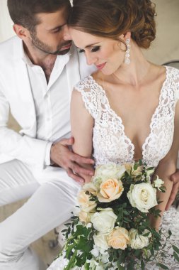 Şık düğün çift damat ve gelin beyaz bir stüdyoda poz. Mutlu çift gülüyor. Beyaz elbise. Beyaz takım. Gençlik. Düğün. Avize. Kanepe. Kapıyı.