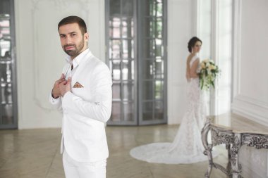 Şık düğün çift damat ve gelin beyaz bir stüdyoda poz. Mutlu çift gülüyor. Beyaz elbise. Beyaz takım. Gençlik. Düğün. Avize. Kanepe. Kapıyı.