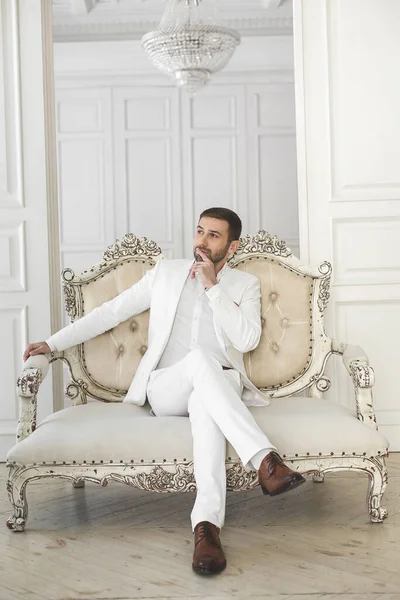 Κομψός νεαρός όμορφος άντρας με γενειάδα σε λευκό κλασσικό κοστούμι. Το εσωτερικό των ξενοδοχείων. Φωτογραφικό στούντιο. Καναπές. Παράθυρο. Καρέκλα. Πολυέλαιο. — Φωτογραφία Αρχείου