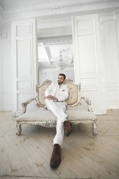 Κομψός νεαρός όμορφος άντρας με γενειάδα σε λευκό κλασσικό κοστούμι. Το εσωτερικό των ξενοδοχείων. Φωτογραφικό στούντιο. Καναπές. Παράθυρο. Καρέκλα. Πολυέλαιο. — Φωτογραφία Αρχείου