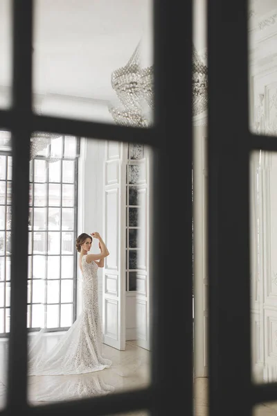 Schöne Braut posiert in Brautkleid in einem weißen Foto Studio. Spiegel. Sofa. Blumenstrauß. Die Tür. Kronleuchter. — Stockfoto