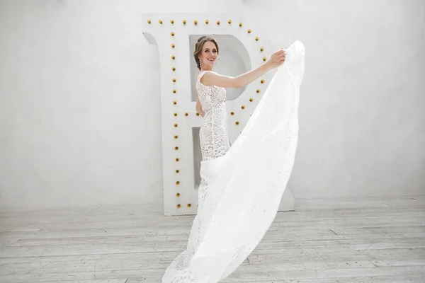 美丽的新娘摆在婚纱礼服在一个白色的摄影棚。镜子.沙发.束.门。吊灯. — 图库照片