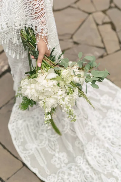 Jemná kytice bílých pivoňky v rukou nevěsty na pozadí vlaku svatebních šatů — Stock fotografie