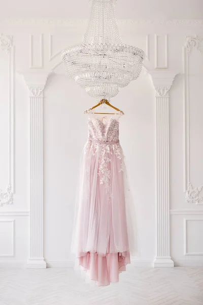 Bohaté růžové svatební šaty visí na lustr v bílém pokoji. — Stock fotografie