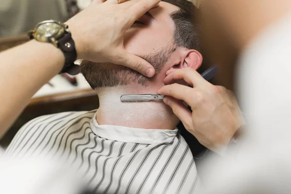 Un giovane è seduto su una sedia in un negozio di barbiere mentre un barbiere si depila la barba. Preparazione dello sposo il giorno del matrimonio — Foto Stock