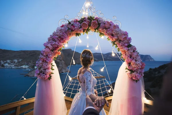 A noiva segura a mão dos noivos contra o fundo de um arco de casamento com luzes brilhantes ao pôr do sol na praia. A cerimônia de casamento — Fotografia de Stock