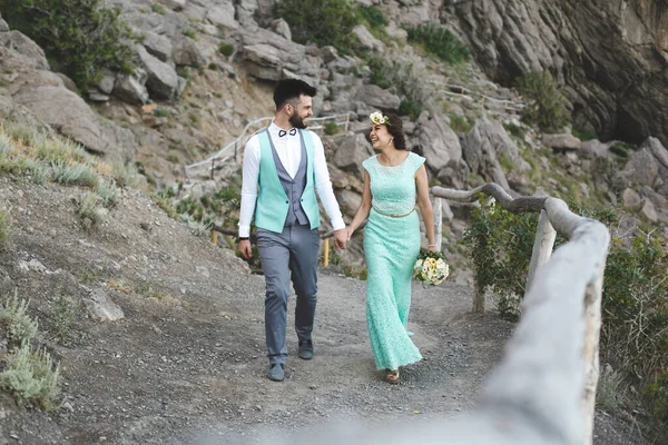 Das Brautpaar über die Natur in den Bergen am Wasser. Anzug und Kleiderfarbe Tiffany. Hand in Hand gehen — Stockfoto