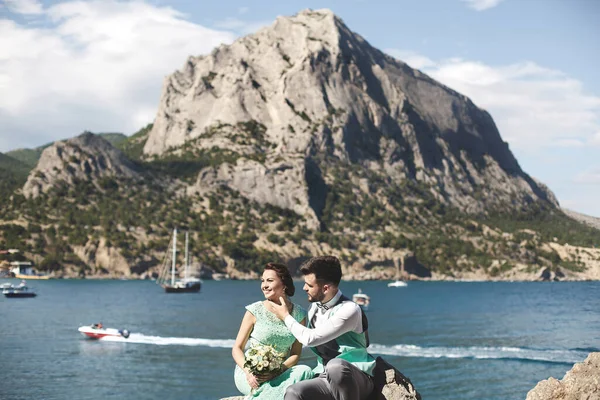 A noiva e o noivo na natureza nas montanhas perto da água. Terno e vestido Tiffany cor. Sentado na pedra — Fotografia de Stock