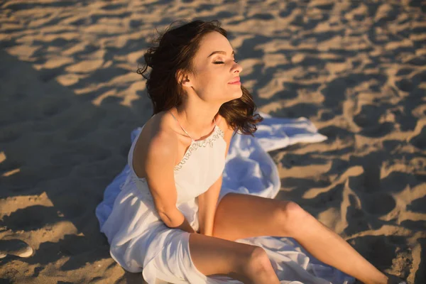 Mooi meisje bruid in een wit jurk zitten op strand zand bij zonsondergang. — Stockfoto