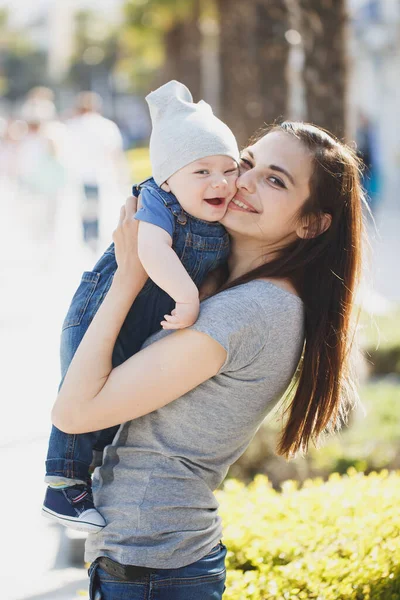 Мила молода матуся цілує щоку красиво одягненого сина і насолоджується приємним сонячним днем. Син на руках матері і посміхаючись — стокове фото