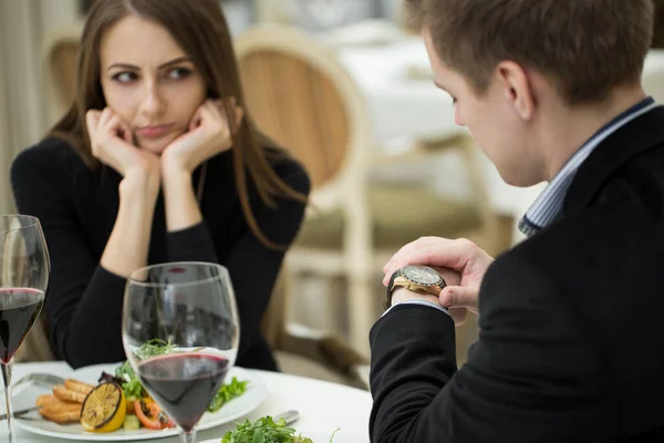 Young woman making an exasperated expression gesture on a bad date at the restaurant. Man looks at his watch.