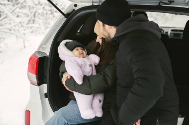Genç, mutlu bir aile: bir adam, bir kadın ve bir bebek kışın karla kaplı bir ormanda bir arabanın bagajında oturuyorlar.