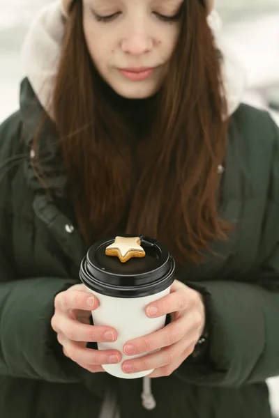 Mujer joven sonriente sostiene una taza de té caliente o café con pan de jengibre en el fondo de un jardín cubierto de nieve — Foto de Stock