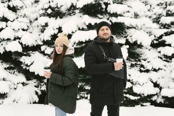 O homem e a mulher têm um resto no parque de inverno. Jovens amigos em roupas de inverno em pé entre árvores nevadas e desfrutando da primeira neve — Fotografia de Stock