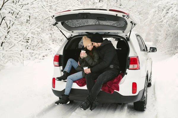 Casal jovem, um homem e uma mulher, estão sentados no porta-malas de um carro em um inverno, floresta nevada, abraçando, beijando e bebendo café — Fotografia de Stock