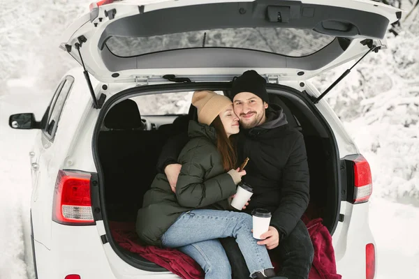 Casal jovem, um homem e uma mulher, estão sentados no porta-malas de um carro em um inverno, floresta nevada, abraçando, beijando e bebendo café — Fotografia de Stock