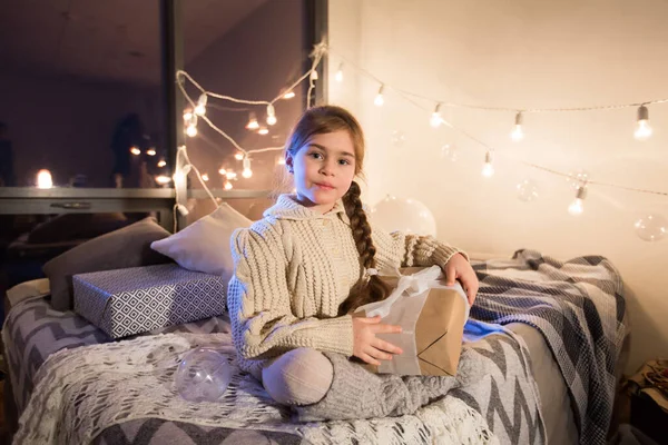 Hermosa niña sentada en la cama con un regalo en sus manos. — Foto de Stock