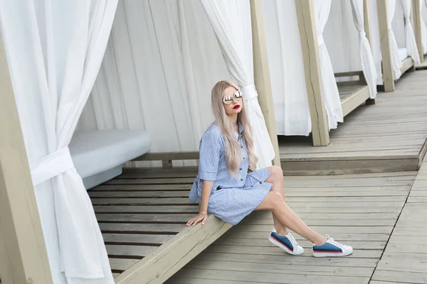 Portret van een mooi blond meisje met lang haar poserend voor de camera op straat. Ze draagt blauwe jurk en een bril.. — Stockfoto