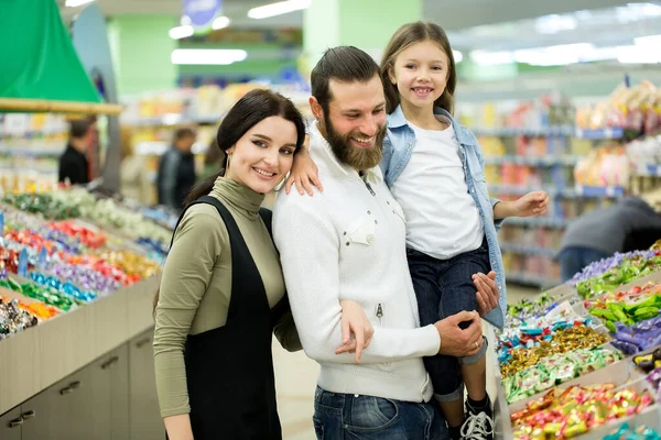 Молодая семья с маленькой девочкой выбирают конфеты и шоколад в большом магазине, супермаркете — стоковое фото