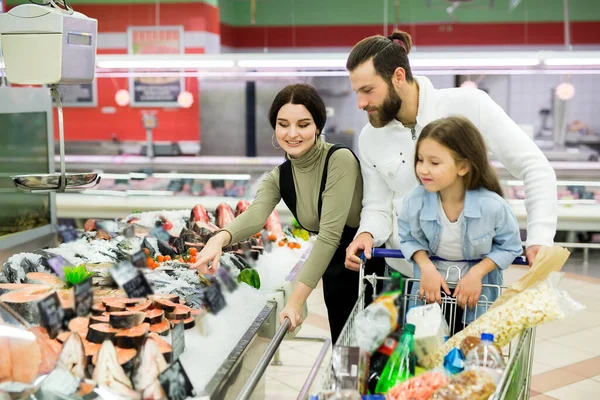 Молодые родители и их милая маленькая дочь улыбаются, выбирая рыбу в супермаркете. — стоковое фото