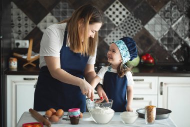 Anne ve kız, un, süt ve modern bir mutfaktaki bir masada oturarak tatlı bir pasta hazırlıyorlar. Elinde çırpı tutan, kasede yumurta karıştıran, annesiyle krep hamuru hazırlayan bir kız.