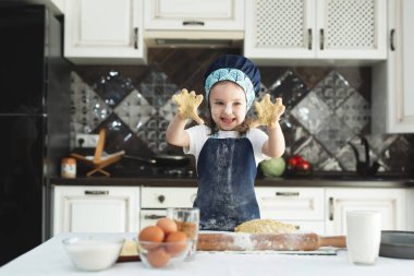 Küçük neşeli kız, elleri önlük ve aşçı şapkasıyla mutfakta hamur yoğurur.