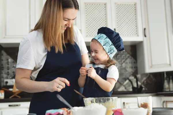 Χαριτωμένο κοριτσάκι και η όμορφη μαμά της σε ασορτί ποδιές και καπέλα παίζουν και γελάνε ενώ ζυμώνουν ζύμη στην κουζίνα — Φωτογραφία Αρχείου
