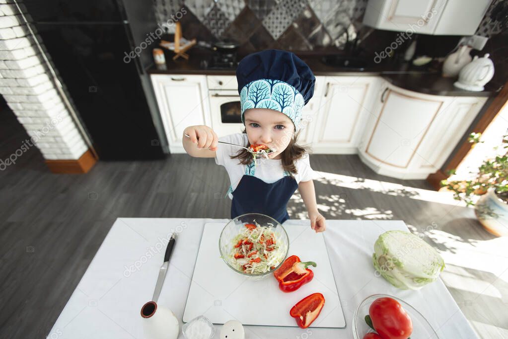 Child in an apron and a chefs hat is eating a vegetable salad in the kitchen and looking at the camera
