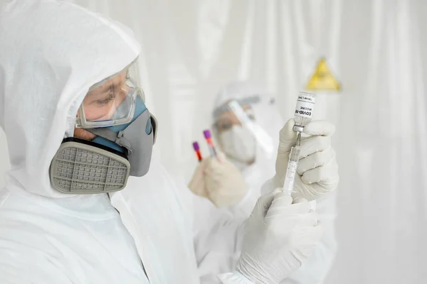 Il medico in tuta protettiva e maschera contiene una siringa per iniezione e il vaccino COVID-19. Scienziati di laboratorio in una tuta che conducono un vaccino di ricerca — Foto Stock