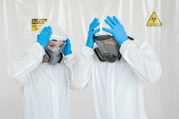 Portrætter af lægen: en mand og en kvinde i beskyttelsesdragter og respiratorer, der holder deres hoveder i karantæne. Covid-19 - Stock-foto