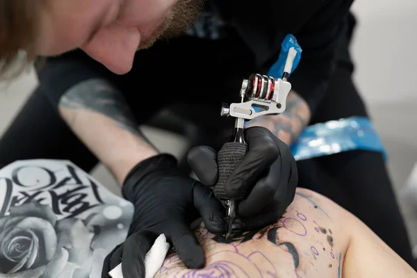 Imagem de perto da tatuagem barbuda artista masculino faz uma tatuagem em uma perna feminina . — Fotografia de Stock