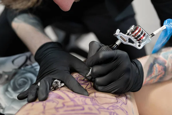 Close up image of the bearded tattoo male artist makes a tattoo on a female leg. — Stock Photo, Image