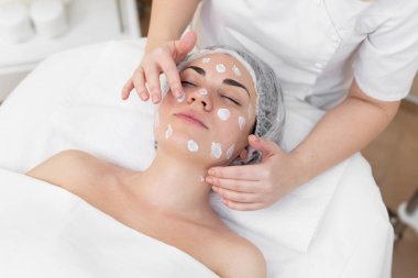 Güzellik uzmanının elleri bir kadının yüzüne krem sürer. Güzellik ve gençlik kavramı. Güzellik uzmanı kızların yüzüne nemlendirici sürüyor.