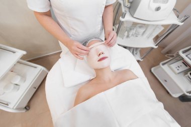 Güzellik uzmanı, kaplıca salonundaki genç bir kızın yüzünden kilden bir maske çıkarıyor. Güzellik uzmanındaki güzel genç kız spa prosedürlerini yapıyor.