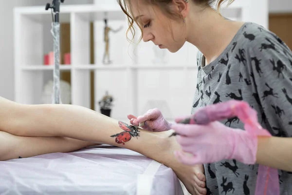 Mestre fazendo tatuagens no estúdio de tatuagem. A tatuadora profissional trabalha em estúdio. Uma mulher de luvas rosa faz uma tatuagem na perna de uma jovem. — Fotografia de Stock