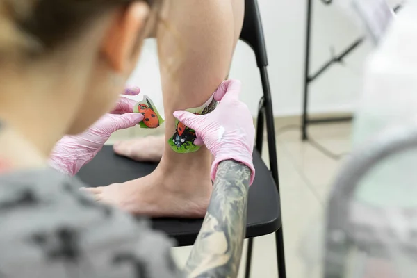 Tattoo artist puts a drawing of a ladybug on the leg of a young woman, the process of creating a tattoo. A girl takes a picture on a womans leg. A tattoo artist makes a tattoo. Close-up — Stock Photo, Image