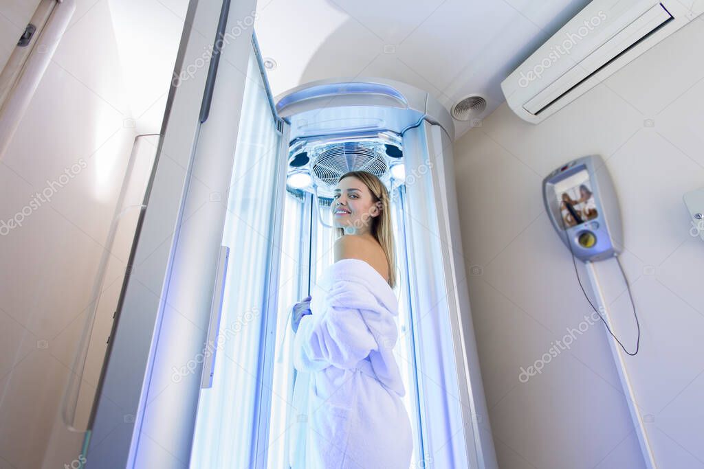 Sexy girl in a white robe is preparing for the tanning procedure in the Solarium. Vertical Solarium. Portrait of a slender girl in a modern Solarium.