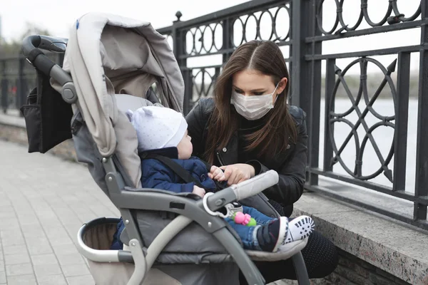 Frau mit Maske auf dem Gesicht zum Schutz vor Coronavirus, covid-19 hält ihr kleines Kind an der Hand, das im Kinderwagen sitzt. — Stockfoto