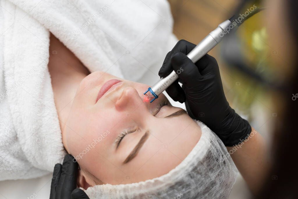 Close-up removal of blood vessels on the face of a diode laser in a cosmetic clinic. Therapist beautician makes a laser treatment to young womans face at beauty SPA clinic.