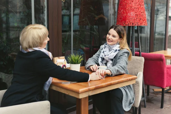 La familia, una madre adulta y su hija están sentadas en una mesa de madera en un café en la calle, mirándose, sonriendo y tomándose de la mano — Foto de Stock